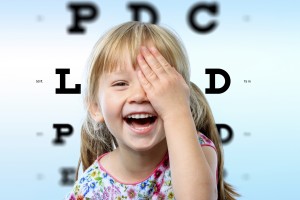 Close up face portrait of happy girl having fun at vision test.Conceptual image with girl closing one eye with hand and block letter eye chart in background.